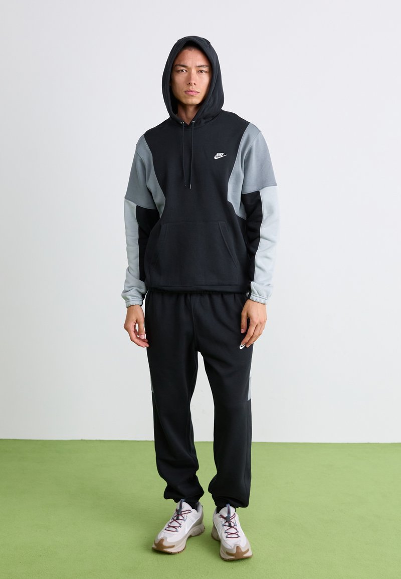 Young man wearing a black and grey Nike hoodie with the hood up and matching black Nike sweatpants and white sneakers, standing on green floor.