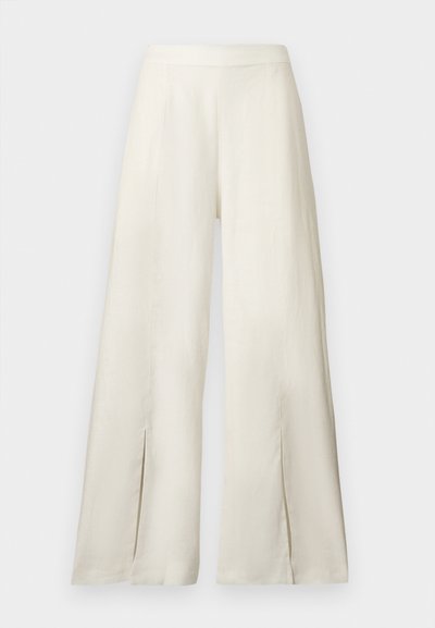 Κρεμ παντελόνια φαρδιάς γραμμής με ψηλή μέση και έντονες σχισμές στο τελείωμα, εκτεθειμένα μπροστά σε απλό φόντο.