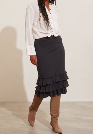 Frau trägt eine weiße, geknöpfte Bluse, die an der Taille gebunden ist, einen schwarzen, mehrlagigen Stufenvolant-Rock und braune kniehohe Wildlederstiefel.