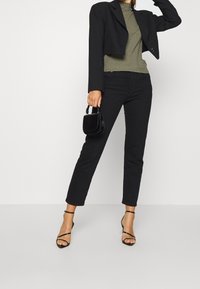 Blazer noir court porté sur un haut côtelé vert, pantalon noir ajusté, et talons noirs à brides ; tenant un petit sac à main noir texturé.