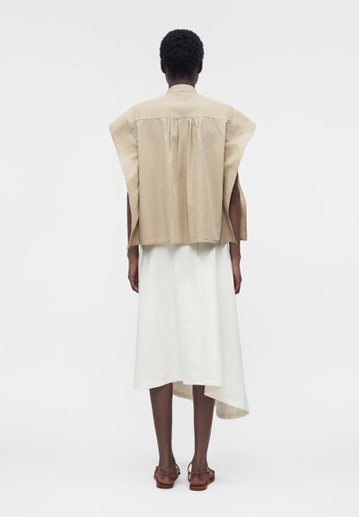 Άτομο με κοντά σγουρά μαλλιά που φοράει μπεζ μαν sleeveless γιλέκο πάνω από λευκό ασύμμετρο φούστα και καφέ σανδάλια, με πλάτη.