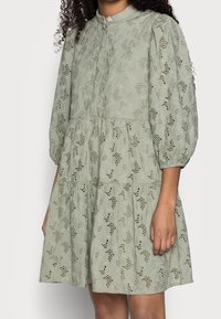 Kortärmad, ljusgrön klänning i texturerad tyg med blommönster och utskärningar, med en lagerdesign och knappstängning.