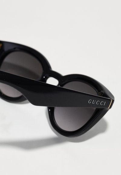 Ochelari de soare negri Gucci cu lentile rotunjite și brațe groase, având logo-ul Gucci pe o braț, pe un fundal alb.