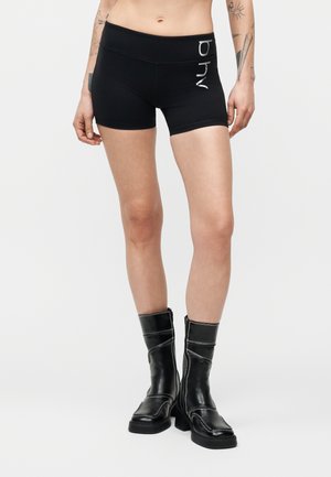 Személy fekete testhezálló rövidnadrágot visel "ohy" felirattal és fekete, középmagasságig érő bőrcsizmát, deréktól a lábfejekig látható, semleges háttér előtt.