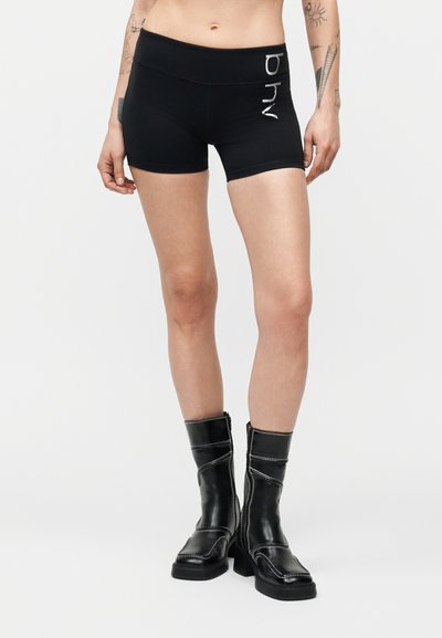 Personne portant un short noir ajusté avec l'inscription « ohy » et des bottes en cuir noires mi-mollet, visible de la taille aux pieds, fond neutre.
