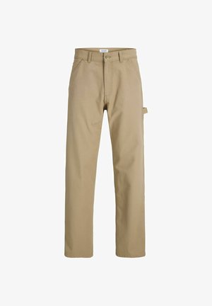 Dreng iført beige jakke, hvid skjorte, kaki bukser, brune sko og hvid kasket, står foran en ensfarvet lys baggrund og smiler.