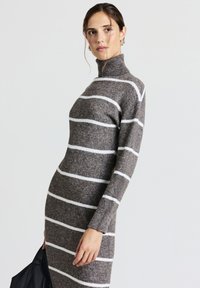 Abito in maglia grigia con strisce bianche, collo alto e maniche lunghe. Il tessuto sembra morbido con una finitura testurizzata. La modella tiene una borsa nera.