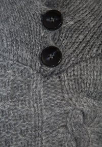 Grå stickad cardigan med ett strukturerat mönster, två runda svarta knappar och mjukt, tjockt garn. Detaljerna inkluderar kabelstickning.