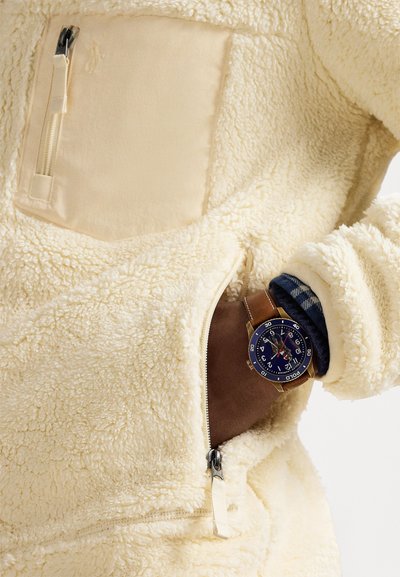 Σκούρο δέρμα χέρι σε τσέπη μπουφάν από φλις κρεμ, φοράει ρολόι με μπλε καντράν και καφέ δερμάτινο λουράκι, και μανσέτα πουκάμισου καρό από κάτω.