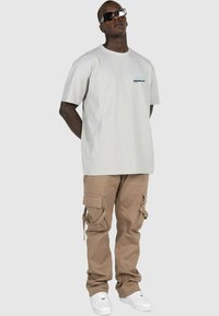 Lys grå t-shirt med lille sort logo på brystet, parret med beige cargo bukser med flere lommer og hvide sneakers.