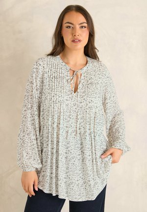 Kvinde iført en løs hvid bluse med små sorte blomsterprint, plisseringer nær halsudskæringen og bindelukning, kombineret med mørke bukser.