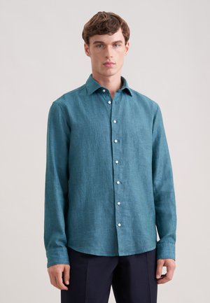 Junger Mann, der ein türkisfarbenes, langärmliges Hemd mit Knöpfen und dunkle Hosen trägt, steht vor einem schlichten, hellen Hintergrund.