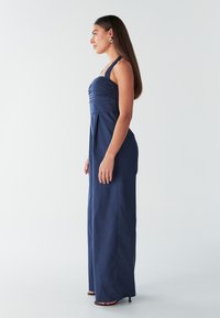 Mornarsko modr kombinezon s prilegajočim zgornjim delom in nabranimi detajli, tekočo tkanino, širokimi naramnicami ter ravnimi hlačnicami, kombiniran s črnimi petkami.