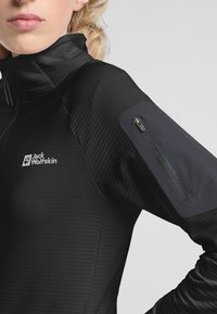 Zwarte performancejas met ribbeltextuur, hoge kraag en een ritszak op de linkermouw. Voorzien van een logo op de borst.