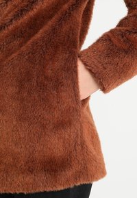 Brun fluffig tyg som visar en hand i en sidoficka, med en mjuk textur och en avslappnad silhuett. Ärmdetaljer synliga vid kanten.