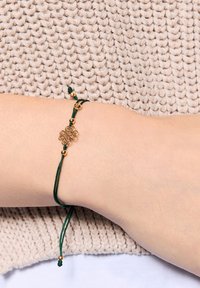 Zielona bransoletka z sznurkiem z dekoracyjnym złotym zapięciem i koralikami, regulowana pod względem rozmiaru, noszona na jasnej skórze nad teksturowanym tłem.