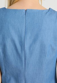 Sininen farkkuhamonen, jossa on neliskulmainen pääntie ja piilotettu takavyö. Kangas on teksturoitu, mikä korostaa siistiä ja yksinkertaista muotoilua.