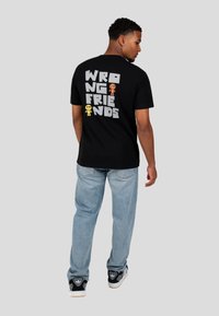 Černé bavlněné tričko s nápisem "WRONG FRIENDS" v tučném šedém písmu na zadní straně, doplněné barevnými ikonami postav. Spárované se světlými džíny.
