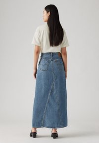 Džinsinė maxi sijonas su aukštu liemeniu, užpakalinėmis kišenėmis, išskirtiniu asimetrišku plyšiu ir šviesiai mėlynu skalbiniu.