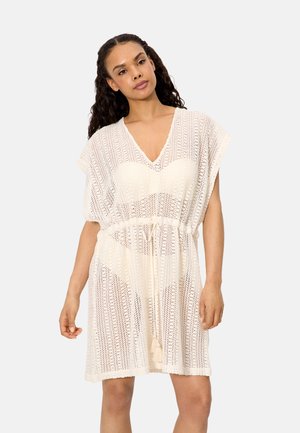 Mujer vistiendo un vestido de punto color crema, de manga corta, con escote en V y cintura con cordón, sobre ropa interior a juego.