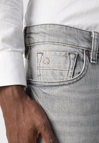 Jeans in denim grigio con una tasca posteriore caratterizzata da un piccolo accento dorato. Fit aderente, con texture e dettagli di cucitura visibili. Sovrapposizione di camicia bianca.