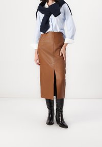 Donna che indossa una camicia azzurro chiaro con bottoni, una gonna midi in pelle marrone con spacco frontale, stivali neri fin sopra il ginocchio e un maglione blu navy appoggiato sulle spalle.
