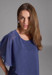 Blouse marine avec des manches transparentes, ornée de sequins près du col, texture lisse et design à encolure arrondie.