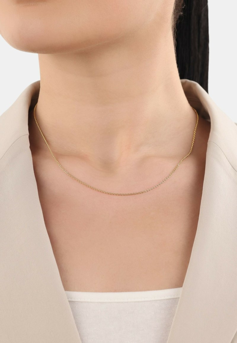Collana di catena d'oro su un collo liscio, con un design delicato caratterizzato da piccoli anelli ovali, abbinata a un blazer beige chiaro.
