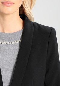 Blazer noir à revers cranté, associé à un pull gris avec un col rond et une décoration en perles au col.