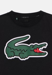 Μαύρο βαμβακερό t-shirt με μεγάλο πράσινο λογότυπο κροκοδείλου με κόκκινες λεπτομέρειες. Λαιμόκοψη τύπου crew και κοντά μανίκια. Λεία υφή.