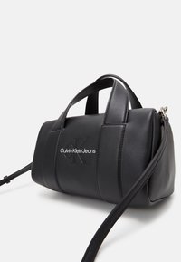 Sac cylindrique noir Calvin Klein Jeans avec double poignées supérieures et une longue bandoulière ajustable sur fond uni.