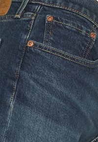 Mörkblå denimjeans med kopparfärgad hårdvara, gyllene sömmar, en texturerad yta och flera fickor placerade fram.