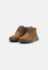Botas de tobillo de ante marrón con punteras redondeadas, cordones en la parte delantera y suelas de goma oscuras. Textura suave con un diseño cosido en la parte superior.