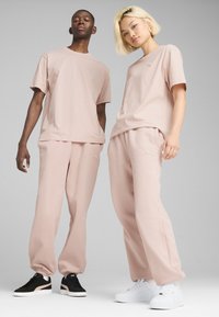 Dwie osoby noszą pasujące jasnoróżowe bawełniane koszulki i spodnie dresowe. Ubrania mają haftowane logo. Obuwie jest różne: czarne i białe.