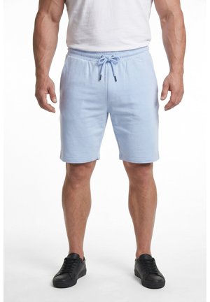 Uomo che indossa pantaloncini azzurri con coulisse, t-shirt bianca e scarpe da ginnastica nere, in piedi con le braccia rilassate lungo i fianchi.