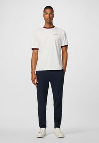 Biele tričko s bordovým rebrovaným golierom a rukávmi, spárované s námorníckymi joggermi so malým logom. Model má na sebe biele tenisky.