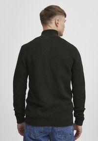 Pulover tricotat de culoare verde închis, cu guler înalt, având mâneci lungi și un tiv ajustat. Textura este vizibilă, cu un model de dungi orizontale.
