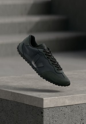 Baskets de sport noires avec semelle texturée et logo, suspendues au-dessus d'un rebord en béton gris avec des escaliers flous à l'arrière-plan.