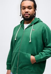 Άνδρας με γενειάδα, φοράει πράσινο φούτερ με φερμουάρ, μπροστινές τσέπες και λευκά κορδόνια, στέκεται μπροστά σε απλό λευκό φόντο.