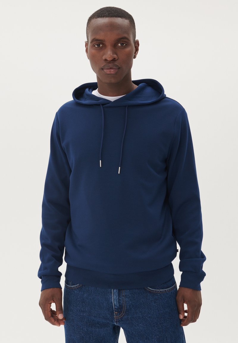 Uomo che indossa una felpa con cappuccio blu navy con lacci e jeans blu, in piedi contro uno sfondo chiaro e semplice.