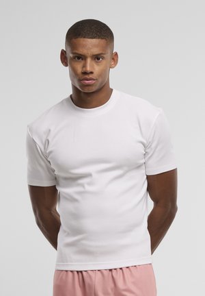 Jeune homme aux cheveux courts portant un t-shirt blanc uni et un short rose clair, debout devant un fond gris neutre.