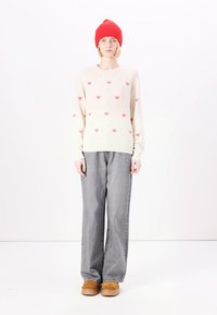 Pull créme avec des motifs de cœurs roses, associé à un jean large gris et des chaussures beiges. Un bonnet en tricot rouge vif complète le look.
