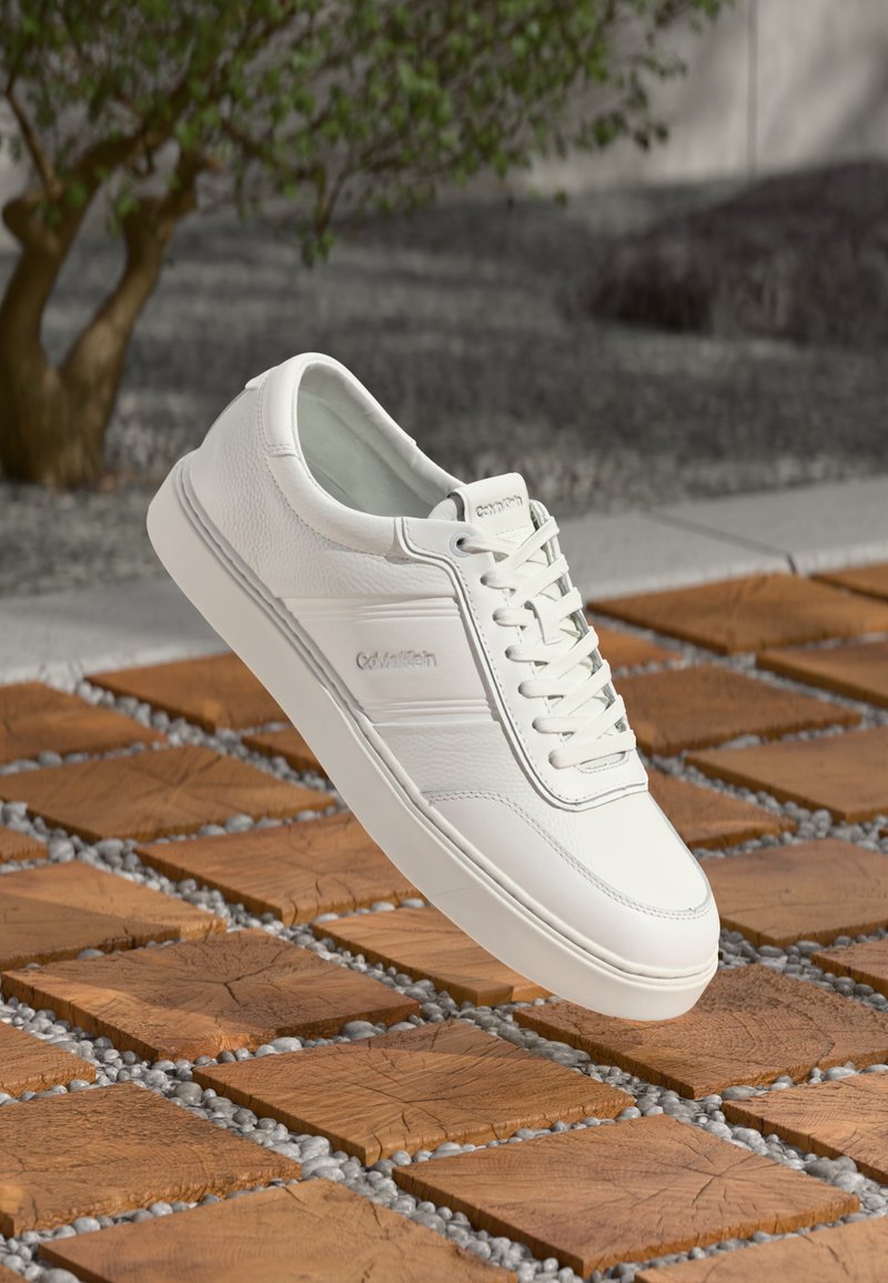 Fehér bőr sneaker sima felülettel, fűzős kialakítással és diszkrét logóval. Minimalista stílust mutat egy mintás csempefelületen.