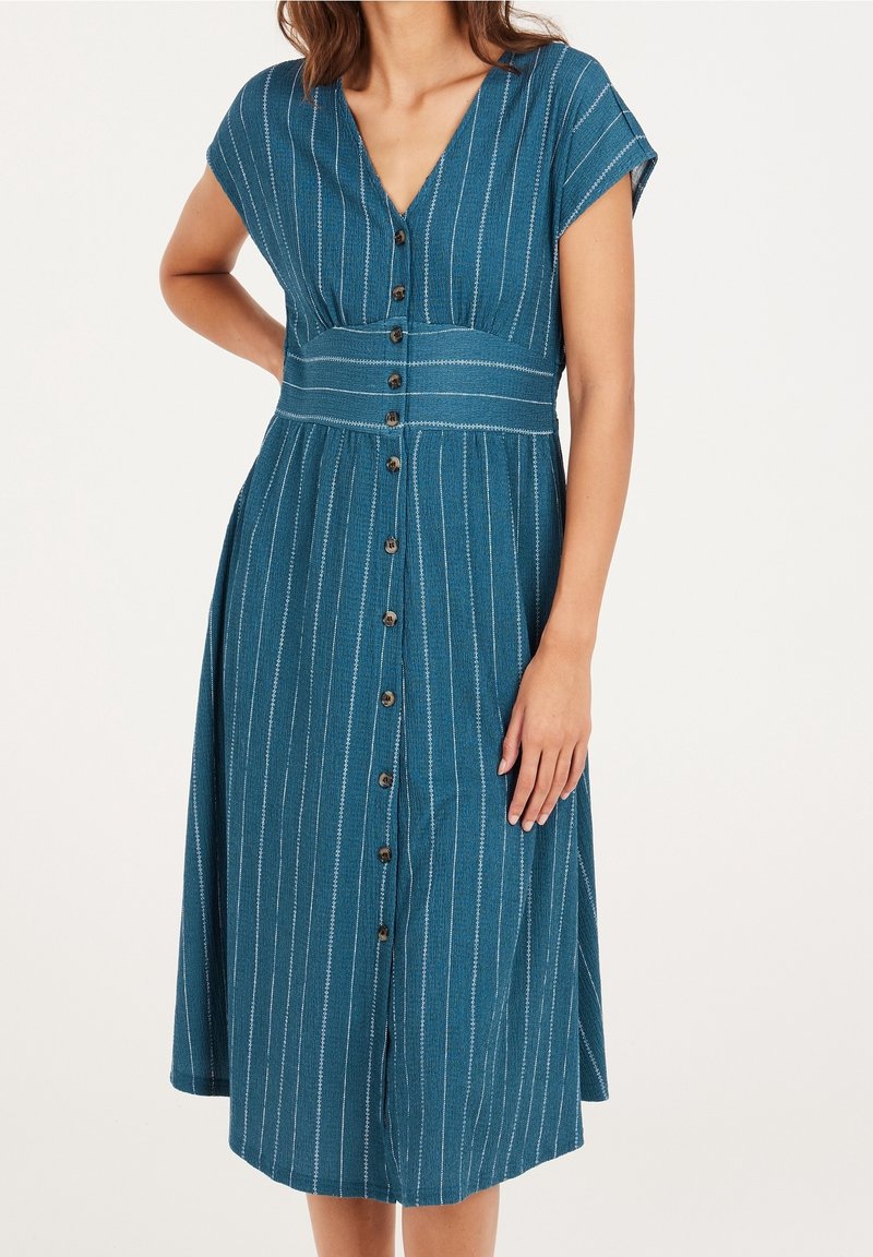 Femme portant une robe midi bleue avec des rayures verticales blanches et des boutons devant, debout avec une main sur la hanche et l'autre le long du corps.
