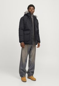 Parka noire avec capuche en fourrure, dotée d'une fermeture éclair et de deux poches avant, portée sur un pull gris et un jean bleu. Chaussures brunes.