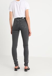 Jeans de mezclilla gris de talle alto, ajustados al cuerpo, con una textura suave, bolsillos traseros y cremallera, combinados con una camiseta blanca y zapatos negros.