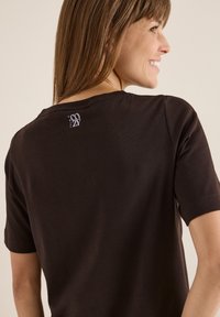 Brązowa bawełniana koszulka z krótkim rękawem, okrągłym dekoltem i małym logo "Calm & Cozy" nadrukowanym z tyłu bluzki w okolicy kołnierza.
