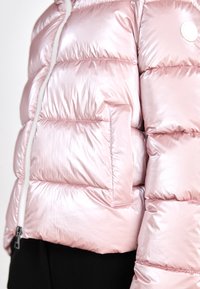 Lyserød dunjakke med et skinnende, reflekterende materiale. Har vandret lynlåsforskæring, en lynlåslomme og en hvid snor ved kraven.