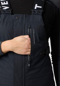Zwarte geïsoleerde overall heeft verstelbare bandjes, een ritszak op de borst en drukknoopsluitingen. Het materiaal heeft een gladde, matte textuur.