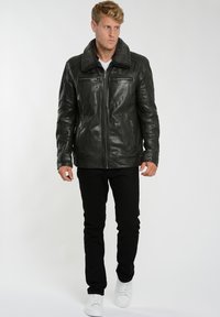 Svart läderjacka med en krage av faux päls, dragkedja framtill, två sidofickor och detaljerad sömnad. Modellen bär svarta byxor och vita sneakers.